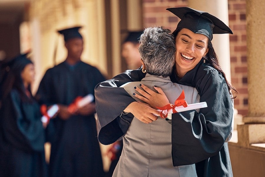 Graduating student in cap and gown hugging an older adult while smiling, holding a diploma..