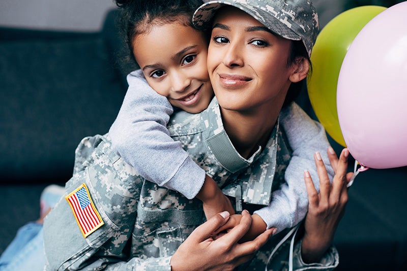 Female soldier smiling with her daughter hugging her from behind, holding colorful balloons.